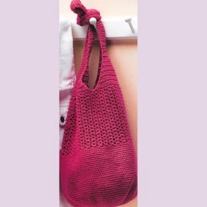 May include: A magenta-colored, crocheted tote bag with a long handle, hanging from a white hook. The bag has a textured, open weave design. The handle is knotted at the top. A white garment hangs to the left of the bag.