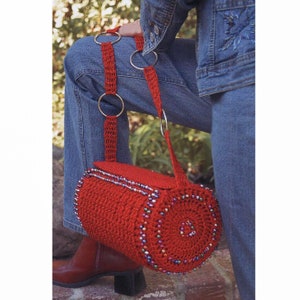 May include: A red crocheted cylinder bag with a beaded trim and a long strap with silver rings. The bag is worn over the shoulder of a person wearing blue jeans.