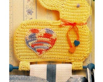 Pode incluir: Um porta-toalhas de coelho de crochê amarelo com um design em forma de coração em vermelho, azul e branco. O coelho tem uma fita laranja e detalhes em forma de coração azul e laranja. Uma toalha listrada em branco e azul pendura abaixo.