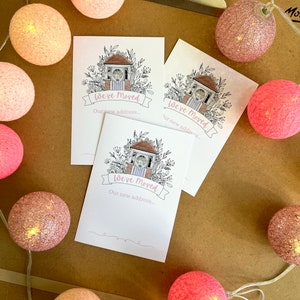 Könnte beinhalten: Drei weiße Karten mit einer Schwarzweißillustration eines Hauses mit einer Uhr auf der Vorderseite. Die Karten haben den Text "We've Moved" und "Our new address..." darauf gedruckt. Die Karten sind von rosa Baumwollkugelleuchten umgeben.