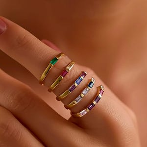 Könnte beinhalten: Eine Sammlung goldener Ringe mit farbigen Baguette-Edelsteinen. Die Ringe zeigen smaragdgrüne, rubinrote, saphirblaue und amethystfarbene Steine. Die Ringe werden an einem Finger präsentiert und zeigen ihre Design- und Farbvariationen.