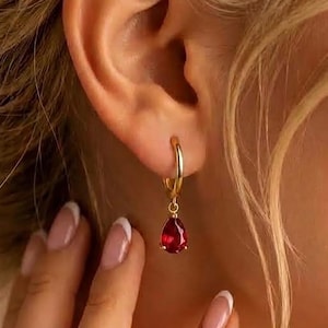 May include: A gold hoop earring featuring a small, dangling, teardrop-shaped red gemstone. The earring is worn in the earlobe, showcasing a polished metallic finish and a vibrant, deep red color of the faceted stone pendant.