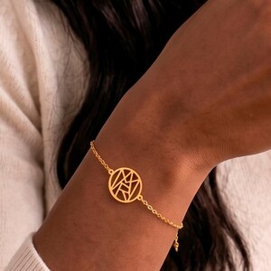 Può includere: Un bracciale a catena color oro con un ciondolo circolare dal design geometrico traforato. Il ciondolo mostra le lettere M, A e R integrate in una cornice circolare. Il bracciale è realizzato con una sottile catena a maglie e una chiusura.