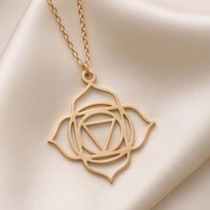 Op de afbeelding: Een goudkleurige ketting met een hanger met een symmetrische, vierbladige bloemvormige omtrek rondom een centrale cirkel en een omgekeerde driehoek. De hanger hangt aan een fijne goudkleurige ketting, gepresenteerd naast een metalen sleutel.
