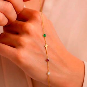 May include: A delicate gold chain bracelet featuring a small gold cross charm and three gemstone accents: green, red, and clear. The bracelet is held up against a soft, neutral background, showcasing its fine details and craftsmanship.