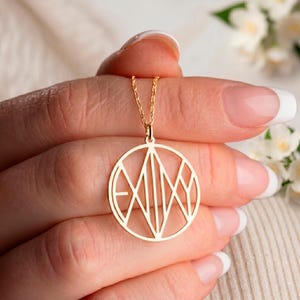 May include: A gold pendant necklace featuring the name "EMILY" in a geometric design within a circular frame. The necklace is held by a person, highlighting the fine chain and the pendant's detailed design. The background is softly blurred.