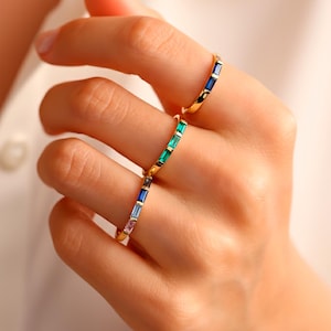 May include: Gold rings adorned with rectangular gemstones in various colours. One ring displays blue stones, another features a gradient of green, blue, and pink gems. The rings are shown on a hand, highlighting the jewellery's design and colour variations.