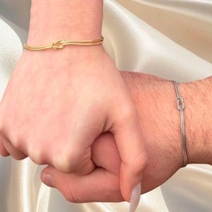 Op de afbeelding: Twee gouden knoop armbanden, één op elke pols. De armbanden zijn eenvoudig en elegant, met een delicaat knoopontwerp.