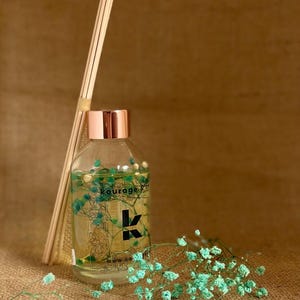May include: A clear glass bottle with a copper coloured lid and a label that reads "kourage" and "www.kourage.co". The bottle is filled with a light yellow liquid and has small green flowers inside. The bottle is sitting on a brown hessian surface with a bunch of small green flowers in the foreground.