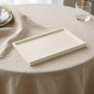 May include: A cream-coloured rectangular serving tray with a raised edge. It sits on a beige linen tablecloth. A glass and a teacup are in the background, suggesting a dining or serving setting.