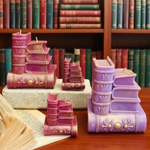 Puede incluir: Una colección de velas con forma de libro en varios tamaños y tonos de morado y burdeos. Las velas están dispuestas sobre una superficie de madera y un bloque de piedra, con una estantería llena de libros al fondo. Cada vela tiene una mecha.