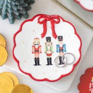 May include: A white ceramic trinket dish with a red scalloped edge and a red bow design. The dish features three nutcracker figures and is decorated with gold stars. Two silver rings rest on the dish.
