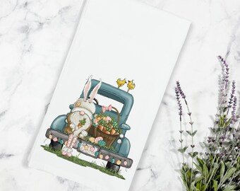 May include: A white bunny with a pink bow sits in the back of a blue pickup truck. The truck is filled with Easter eggs and a basket of carrots. The truck has a license plate that says "Easter".