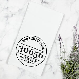 May include: White kitchen towel with a black circle design that says "Home Sweet Home" with the number "30656" and "Between - GA" printed on it.