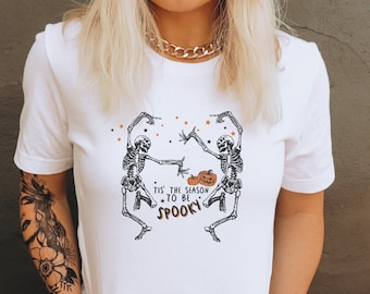 May include: White t-shirt featuring a graphic of two skeletons dancing and holding pumpkins. The text on the shirt reads "Tis the season to be spooky."