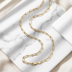 May include: A gold-toned paperclip chain necklace featuring elongated rectangular links with a polished finish. The necklace includes a secure lobster claw clasp mechanism at the end, showcasing a consistent metallic yellow hue throughout the entire piece.