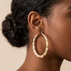 May include: A large, circular hoop earring crafted from gold-toned metal, featuring a textured design that mimics the segmented appearance of bamboo stalks. The earring is worn in the earlobe, showcasing a polished, reflective surface finish.