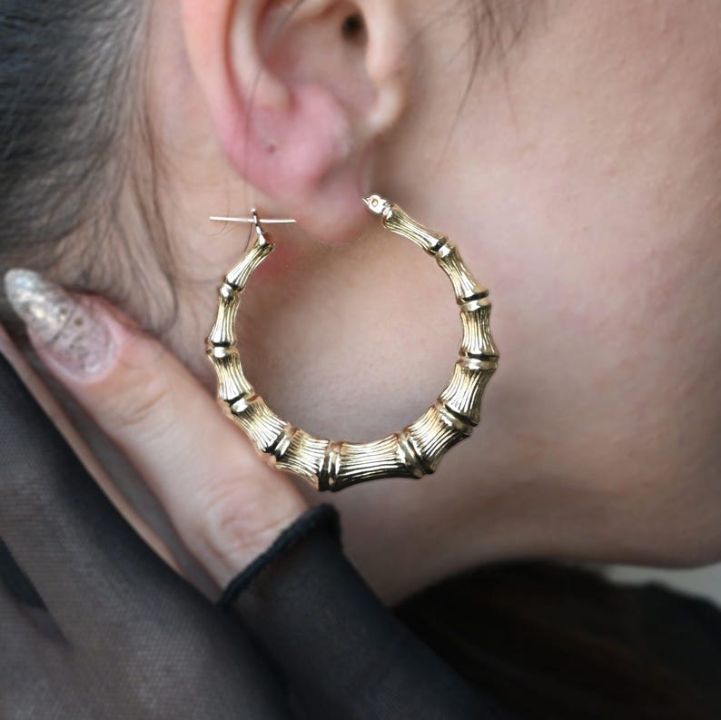 A woman's hand holds a large gold bamboo-style hoop earring to her ear, showing the earring's textured surface and size.