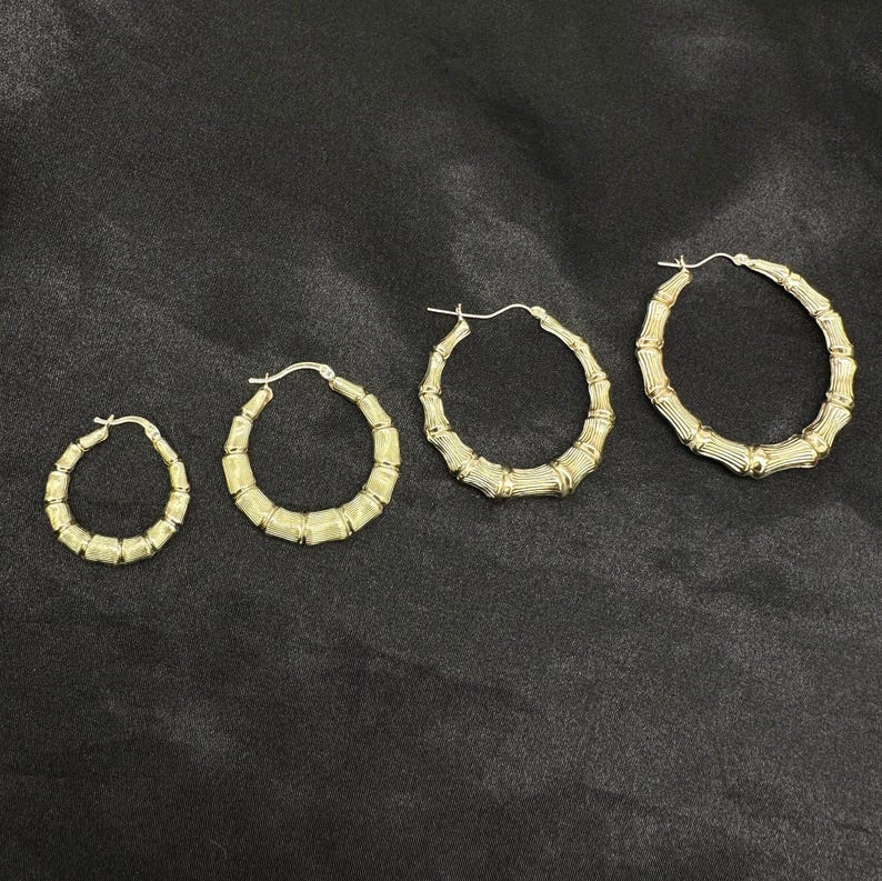 Four gold bamboo-style hoop earrings of increasing size are laid out in a row on a black fabric surface.