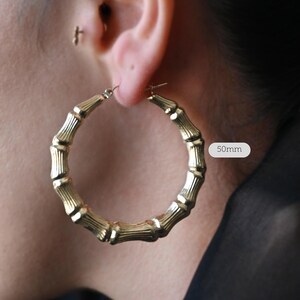 A woman's ear with a large gold bamboo-style hoop earring, with a white text overlay indicating "50mm".
