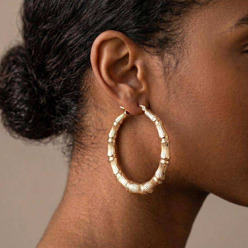 May include: A large, circular hoop earring crafted from polished gold-toned metal, featuring a textured design that mimics the segmented appearance of bamboo stalks. The earring is worn in the earlobe, showcasing a shiny, reflective surface.