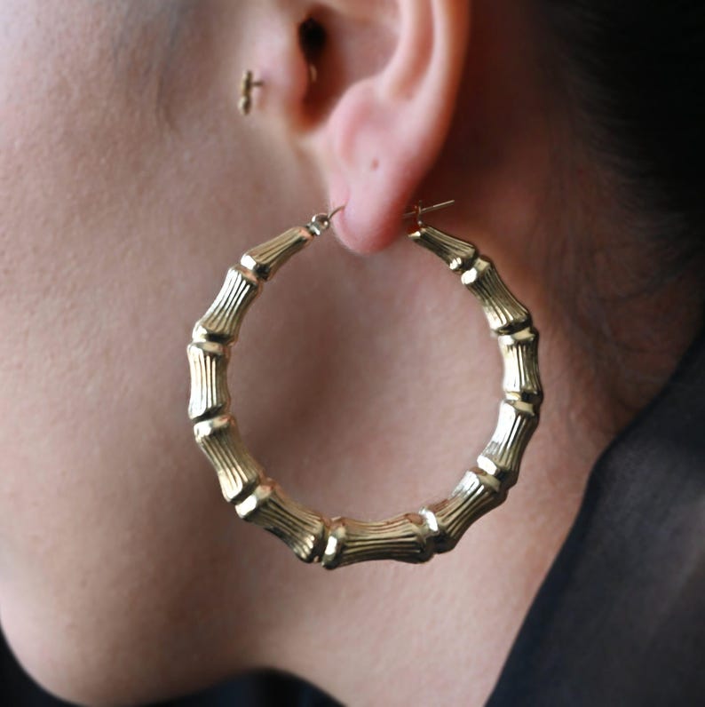 A woman's ear wearing a large, gold, bamboo-style hoop earring, with a small stud earring visible higher on the ear.