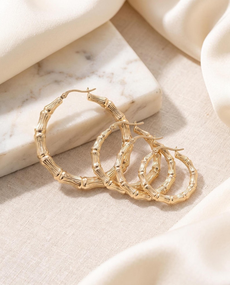 Three gold bamboo-style hoop earrings of different sizes are arranged on a light-colored fabric and marble surface.