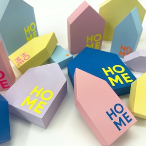Könnte beinhalten: Eine Sammlung von hausförmigen Holzblöcken in verschiedenen Pastell- und kräftigen Farben wie Blau, Rosa, Gelb und Lavendel. Jeder Block trägt das Wort HOME in Kontrastfarben, und einige Blöcke sind mit kleinen Herzsymbolen versehen.