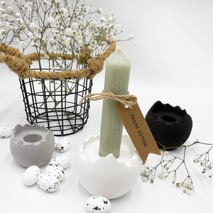Könnte beinhalten: Eine Osterdekoration mit einem schwarzen Drahtkorb mit weißen Blumen, gesprenkelten Eiern und Kerzenhaltern. Eine grüne Kerze befindet sich in einem weißen eiförmigen Halter, mit einem Etikett "Frohe Ostern".