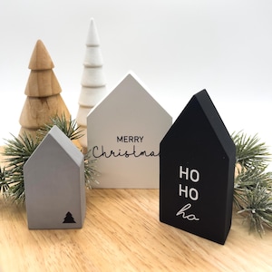 Könnte beinhalten: Drei Holzhäuser mit weihnachtlichem Text. Das weiße Haus sagt "Frohe Weihnachten", das schwarze Haus sagt "Ho Ho Ha" und das graue Haus hat einen kleinen Weihnachtsbaum darauf.