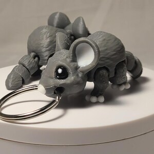 May include: A gray 3D-printed mouse keychain with a silver key ring. The mouse has large black eyes, white ears, and white paws. The mouse is articulated and has a detailed, textured surface.