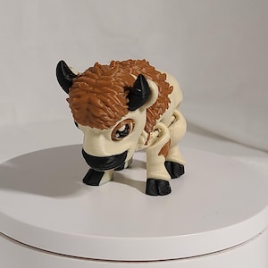 May include: A small, white and brown bison figurine with black hooves and horns. The figurine has a furry texture and is posed in a sitting position.