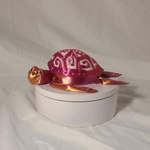 May include: A decorative turtle figurine in shades of pink and gold. The turtle's shell features a patterned design, and it sits atop a white, round base. The figurine is likely a decorative item.