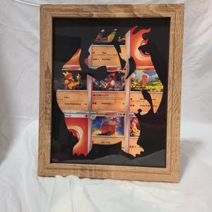 May include: A framed artwork featuring Pokémon trading cards. The cards are arranged within a silhouette of a Charizard, with a wooden frame. The cards display various Pokémon characters in vibrant colors, set against a black background.