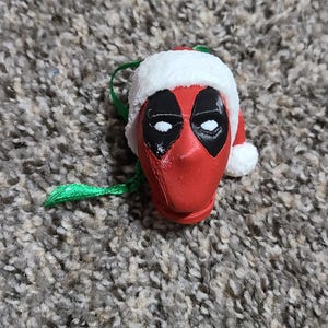 May include: A red Deadpool head Christmas ornament with a white Santa hat and green string. The ornament features black eye shapes and a white trim on the hat. The ornament is a festive decoration.