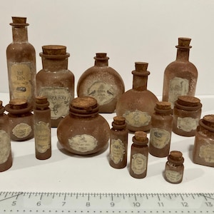 May include: A collection of 14 small glass bottles with cork stoppers. The bottles are various shades of brown and have labels with different text, including "Bat Tails", "Dragon Horn", and "Raven Down".