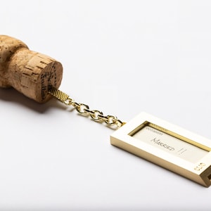 Op de afbeelding: Een gouden metalen sleutelhanger met een champagne kurk eraan bevestigd. De sleutelhanger heeft een klein rechthoekig frame met het woord "Getrouwd" erin.