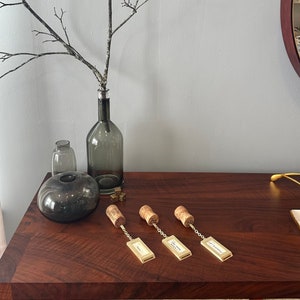 Op de afbeelding: Vier gouden flessenstoppers met kurken en een ketting die aan een rechthoekig label is bevestigd. De labels hebben de woorden "Happy Hour", "Wine Time", "Cheers" en "Let's Get Tipsy" erop geschreven. De stoppers liggen op een houten oppervlak.