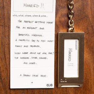 Op de afbeelding: Een witte kaart met handgeschreven tekst die luidt: "occasion MARRIED!! who, what, where, when & wine... THE PERFECT SETTING MADE FOR AN ELEGANT AND BEAUTIFUL WEDDING. A MAJESTIC DAY IN NYC WITH FAMILY AND FRIENDS, WHAT MORE COULD WE ASK FOR? WE MARRIED, DINED, DANCED, AND LOVED. A DREAM COME TRUE. CLIO" Een gouden metalen sleutelhanger met een klein wit kaartje waarop staat: "occasion MARRIED!! CLIO"