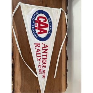 May include: White pennant with the text "CAA MOTOR LEAGUE ONTARIO" in a blue and red oval, and "ANTIQUE AUTO RALLY-CNE" in red. The pennant has white ties and is displayed on a wooden surface.