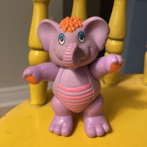Könnte beinhalten: Eine violette Elefantenfigur mit großen Ohren, einem erhobenen Rüssel und orangefarbenem Haar auf dem Kopf. Die Figur hat orangefarbene Akzente an Füßen und Händen sowie einen rosa gestreiften Bauch. Sie hat große, ausdrucksstarke Augen.