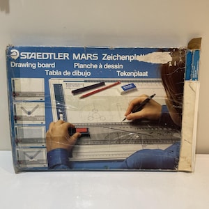 May include: A Staedtler Mars drawing board box featuring a blue and white design with text in multiple languages. The packaging displays a graphic of a person using the board with a drafting tool, a red pencil, and a white eraser.