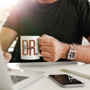 Peut inclure: Une tasse en c&eacute;ramique blanche avec le texte "DR. Robinson" en lettres brunes et orange. La tasse est tenue par une personne portant un t-shirt noir et une montre en cuir marron.