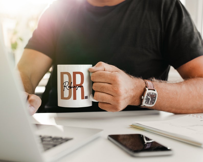 Peut inclure: Une tasse en c&eacute;ramique blanche avec le texte "DR. Robinson" en lettres brunes et orange. La tasse est tenue par une personne portant un t-shirt noir et une montre en cuir marron.