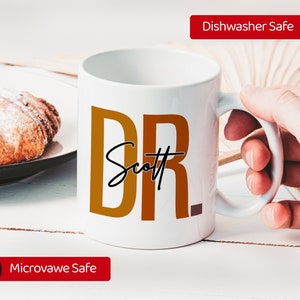 Peut inclure: Une tasse en c&eacute;ramique blanche avec un motif brun et orange qui dit "DR Scott". La tasse est lavable au lave-vaisselle et au micro-ondes.