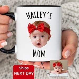 May include: White ceramic mug with a black interior and handle. The mug features the text "HAILEY'S MOM" and a photo of a baby wearing a red bow. A smaller image of the baby is also present.