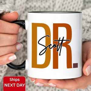 Peut inclure: Une tasse en c&eacute;ramique blanche avec un bord et une poign&eacute;e noirs. La tasse porte le texte "DR" en orange et marron avec le nom "Scott" &eacute;crit en cursive en dessous.