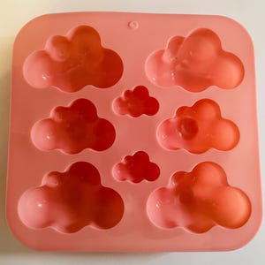 Puede incluir: Un molde de silicona rosa con cavidades en forma de nube. El molde contiene varios objetos en forma de nube en tonos naranja y rojo. El molde es cuadrado con esquinas redondeadas y un pequeño agujero en la parte superior.