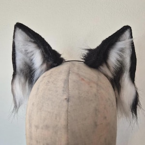 Op de afbeelding: Een paar wolfsoren van imitatiebont in zwart en wit, bevestigd aan een dunne hoofdband. De oren staan rechtop en hebben een realistisch ontwerp met een mix van zwart en wit bont, wat een opvallend contrast creëert. Ontworpen als kostuumaccessoire.