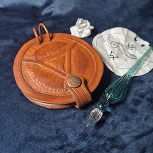 Puede incluir: Un estuche redondo de cuero color canela con un diseño triangular en relieve y un cierre de broche metálico, que presenta dos pequeñas presillas en la parte superior. Junto a él hay una pluma de cristal translúcido color verde azulado.